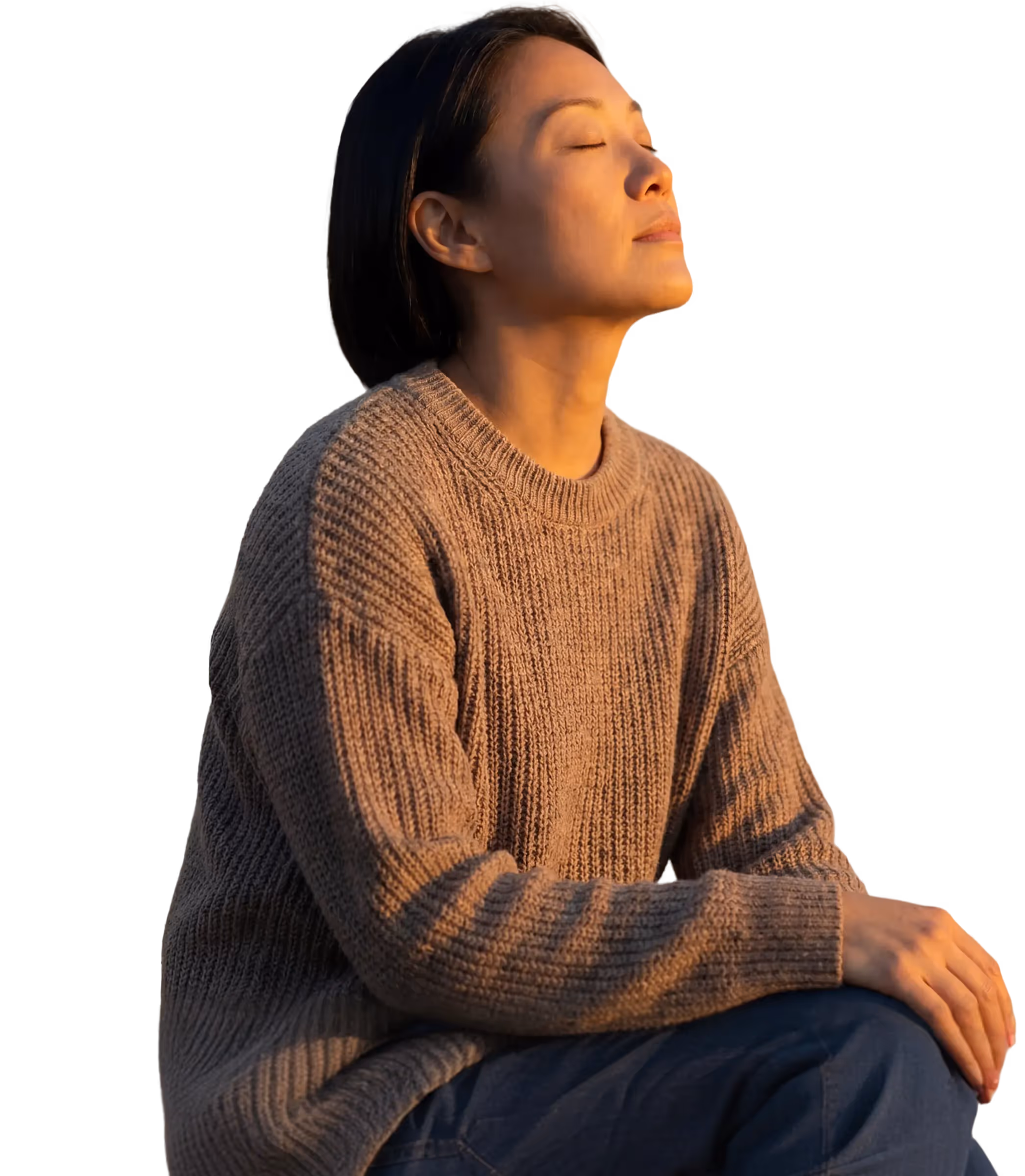 Woman with closed eyes enjoying sunlight while wearing a brown knit sweater and sitting with hands on knee.